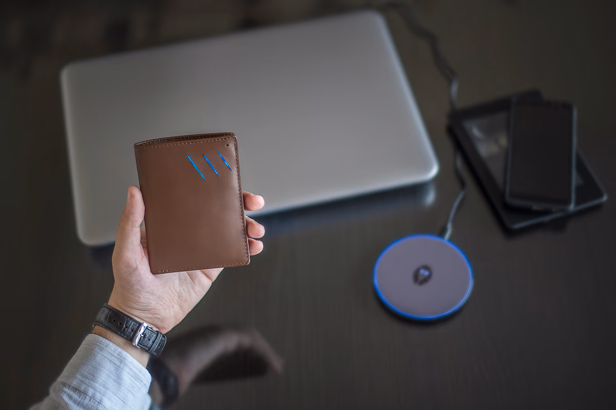 A hand holding a wallet in front of a laptop and smart devices on a desk